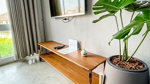 A modern room with a wooden desk, a potted plant, a flat-screen TV mounted on the wall, and a large window with curtains. - Bedgasm Hostel Chanthaburi (Chanthaburi)