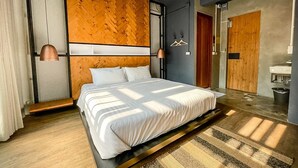 A modern bedroom with a large bed, a wooden accent wall, and a view of the bathroom. - Bedgasm Hostel Chanthaburi (Chanthaburi)