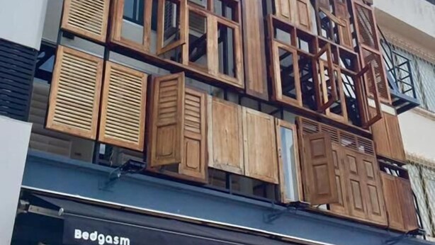 A building with multiple wooden shutters and windows, a sign reading "Bedgasm" below the entrance, and a balcony on the right side. - Bedgasm Hostel Chanthaburi (Chanthaburi)