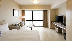 Quarto de hotel com uma cama grande, uma escrivaninha com televisão, uma mesinha e uma janela com cortinas.