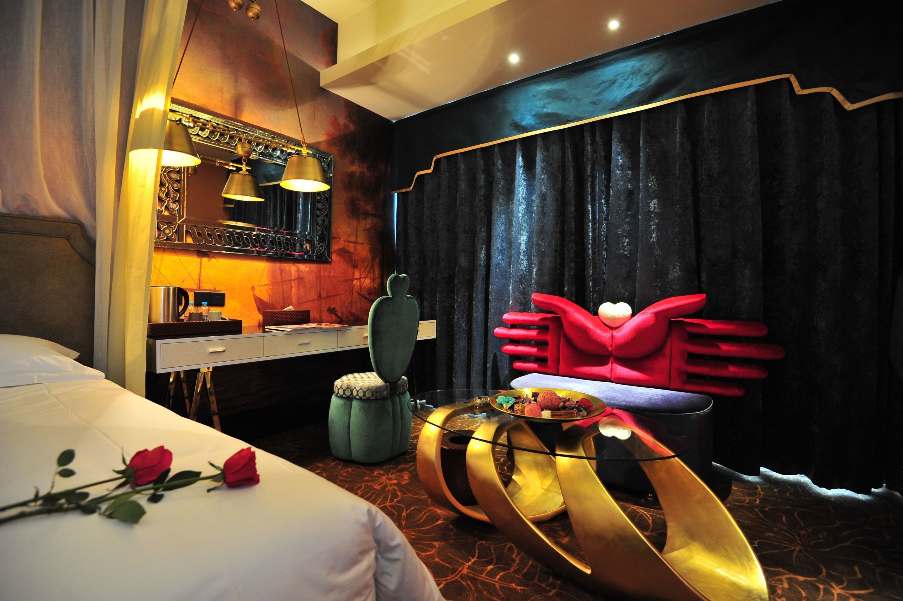 A modern hotel room with a bed, a vanity with a mirror, a green chair, a red armchair, and a unique gold coffee table.