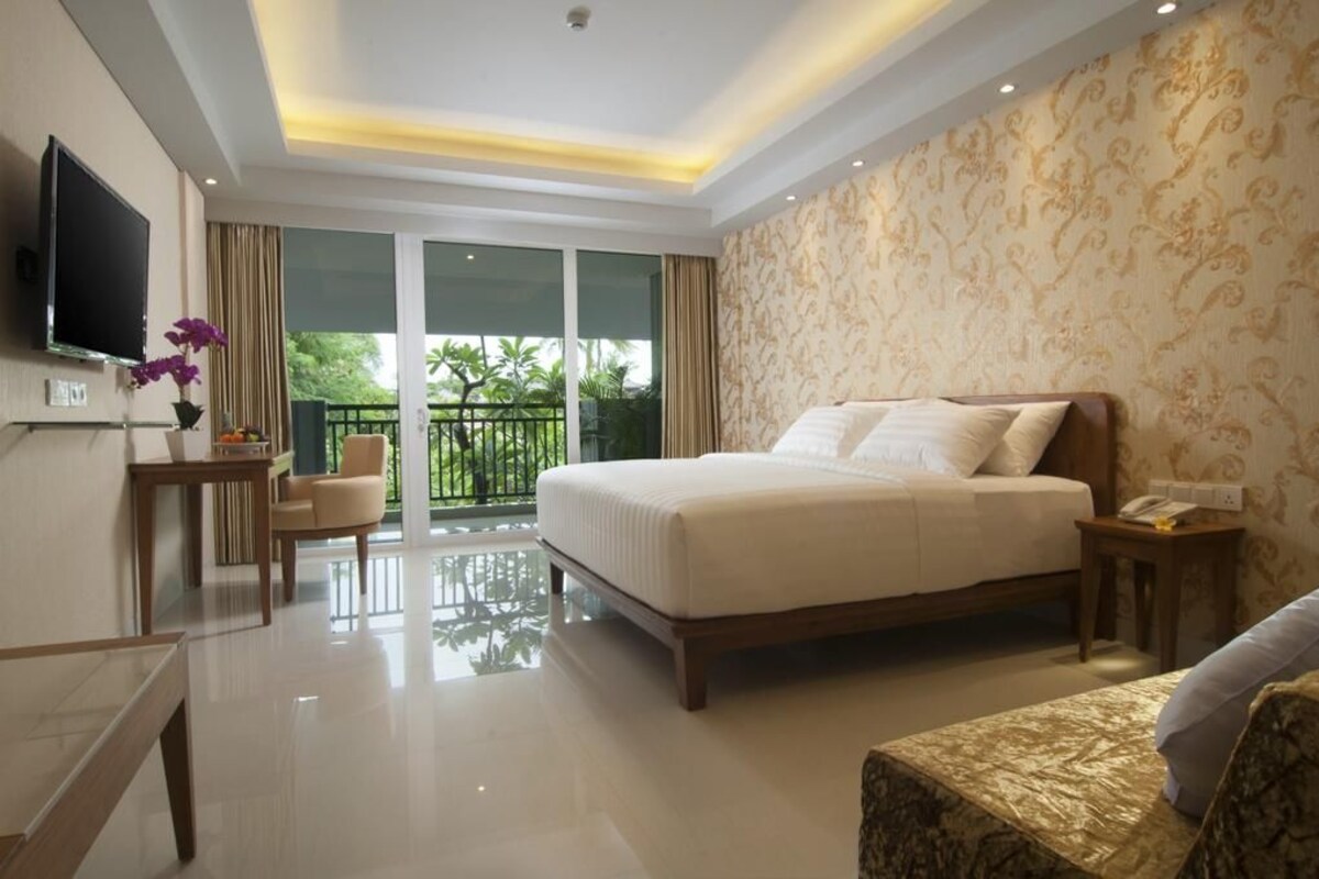 A hotel room with a large bed, a TV, a desk with a chair, a small table, and a balcony with a view of greenery.