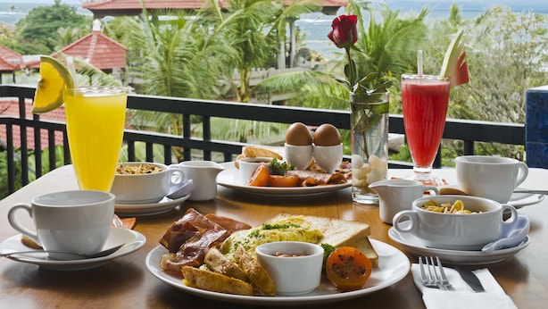 Una mesa de desayuno con diversos platos y bebidas, con vistas a un paisaje con palmeras y un edificio de techo rojo.