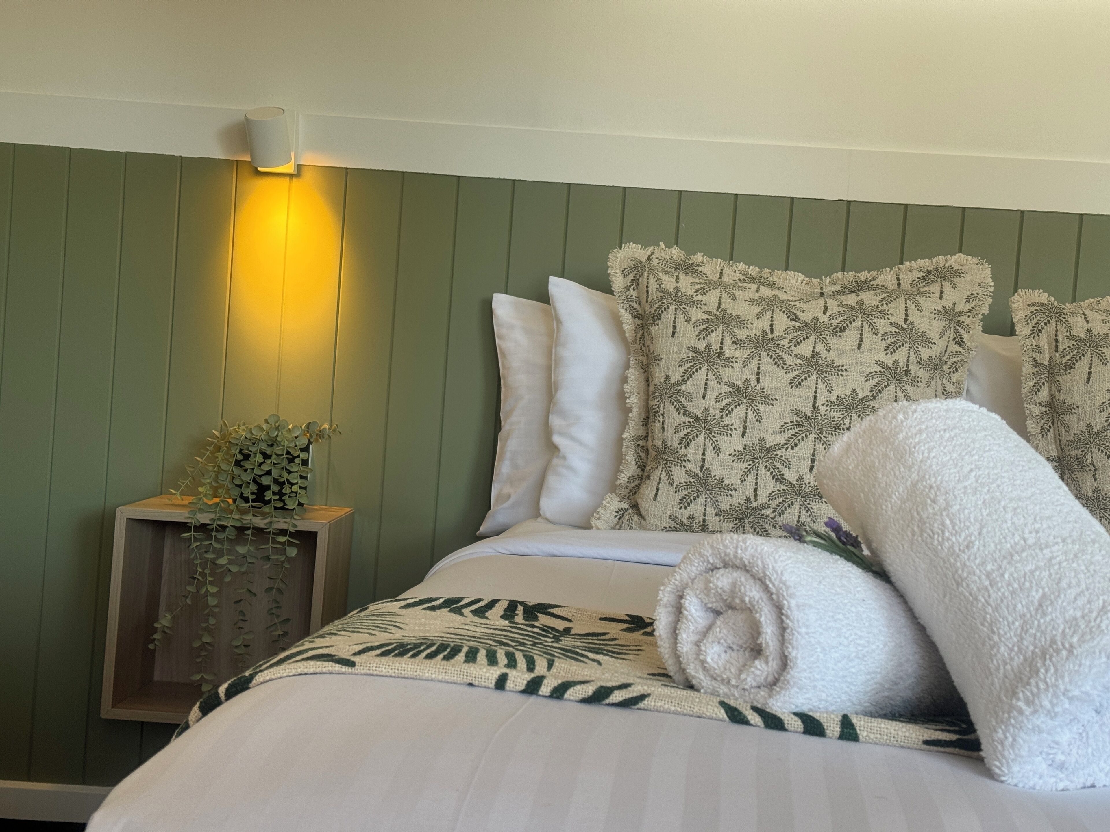 A neatly made bed with white linens and a palm leaf pattern, a bedside lamp, and a small wooden nightstand with a plant.
