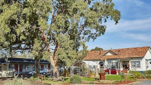 A two-story house with a red roof, a covered porch, and a garden with various plants and a small tree. - The Highlander Motel - 24HR Check In (Maryborough)