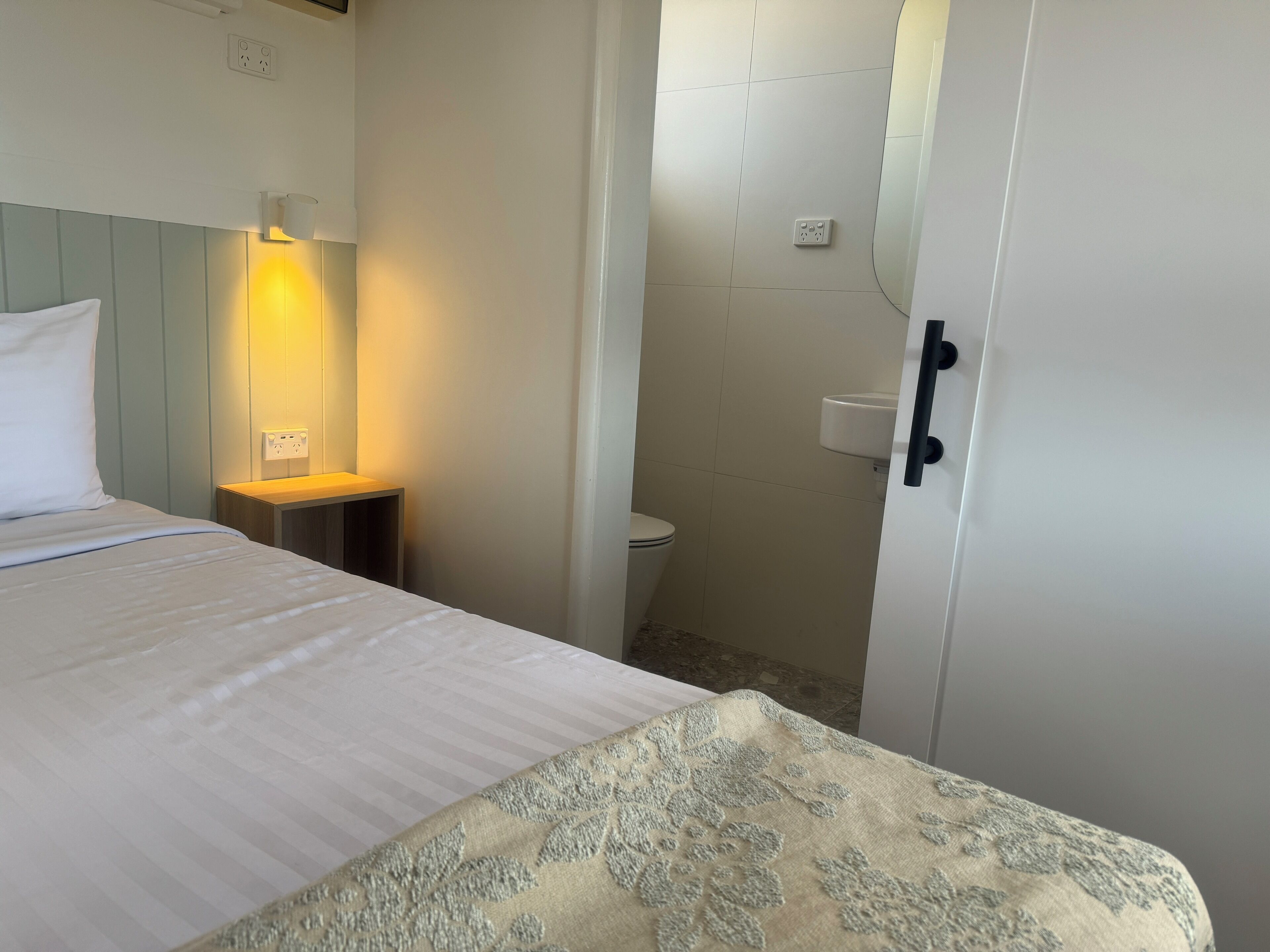 Una habitación de hotel con cama, mesita de noche con lámpara, inodoro y puerta.