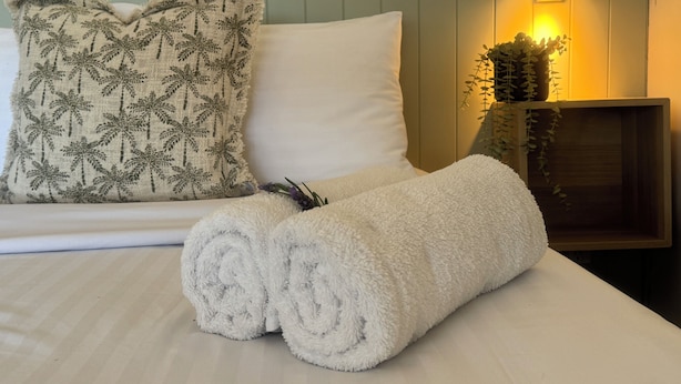 A neatly made bed with a rolled white towel, a pillow with a palm tree pattern, and a bedside lamp with a plant. - The Highlander Motel - 24HR Check In (Maryborough)