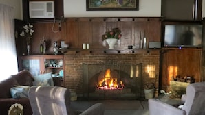 A living room with a brick fireplace, two grey armchairs, a sofa, and a television on a wooden stand. - The Highlander Motel - 24HR Check In (Maryborough)