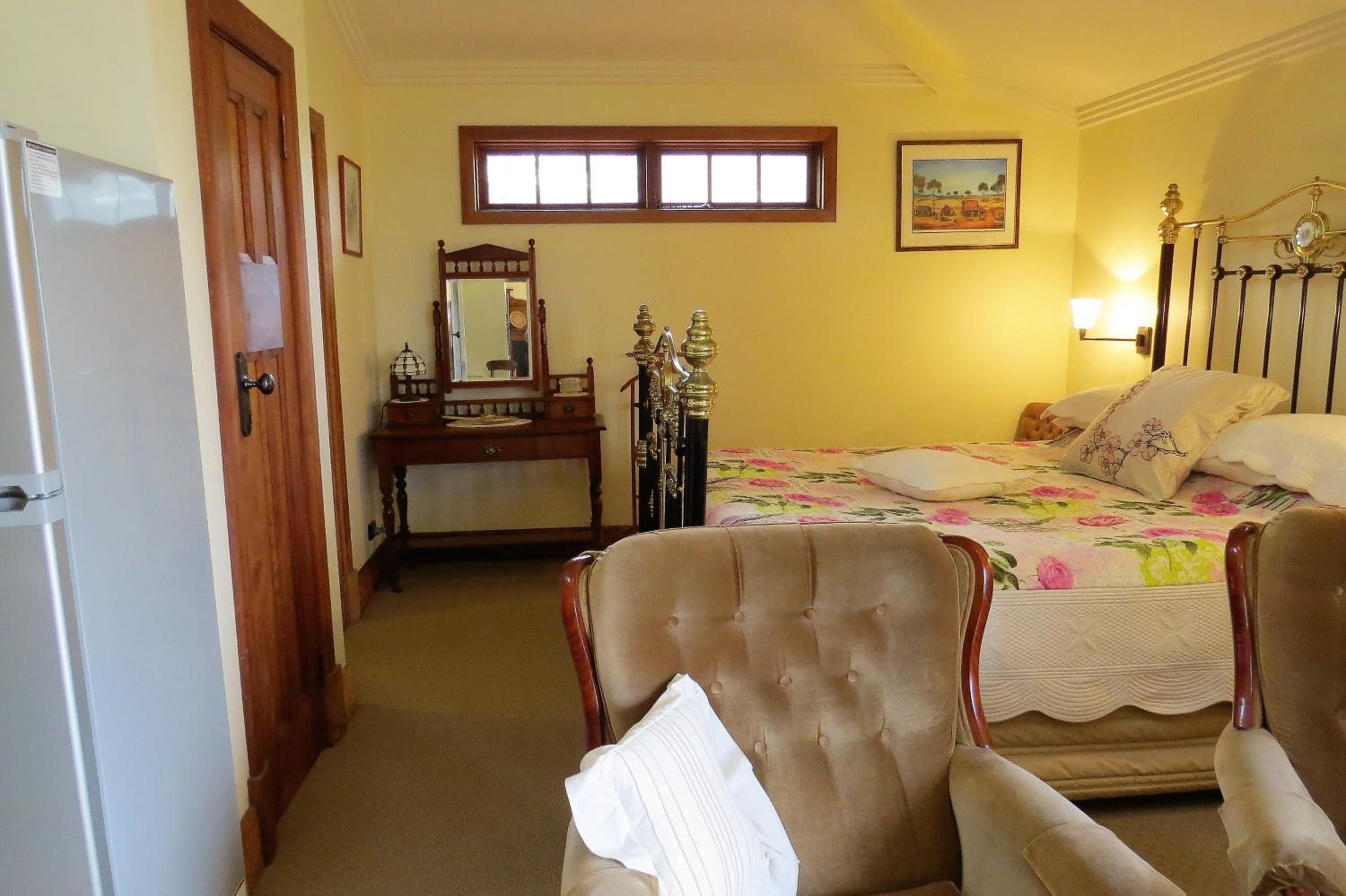 A bedroom with a bed, two armchairs, a dresser with a mirror, a window, and a painting on the wall.