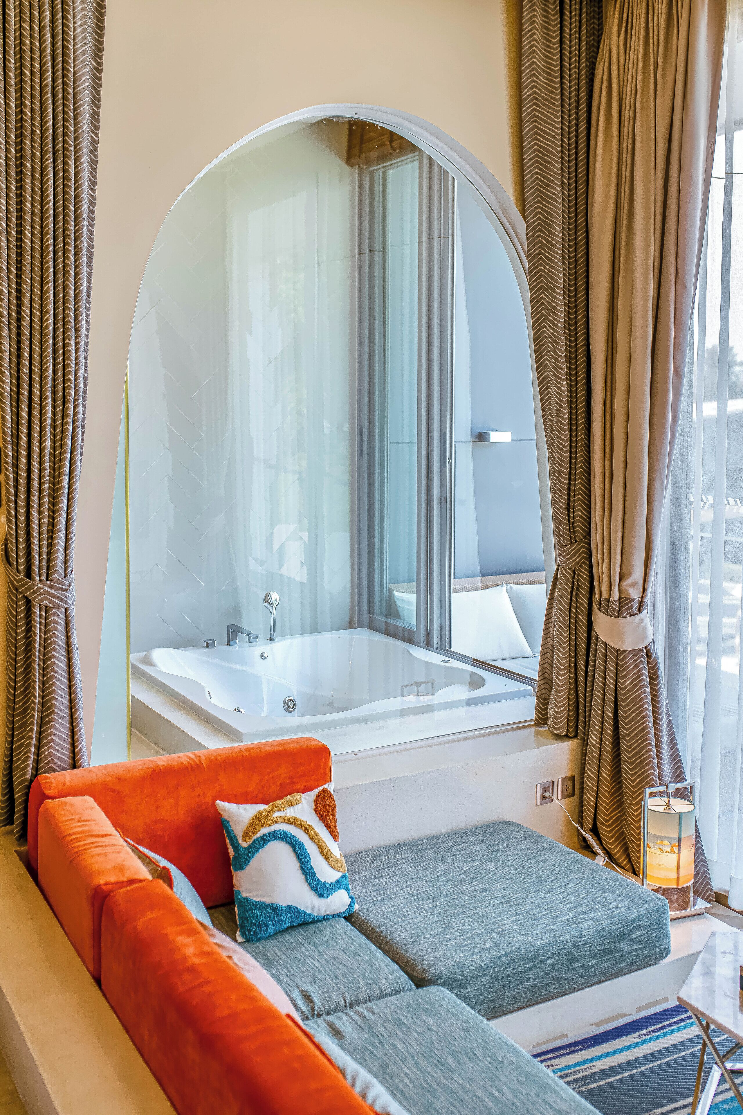 A modern bathroom with a large mirror, a bathtub, and a seating area with cushions.