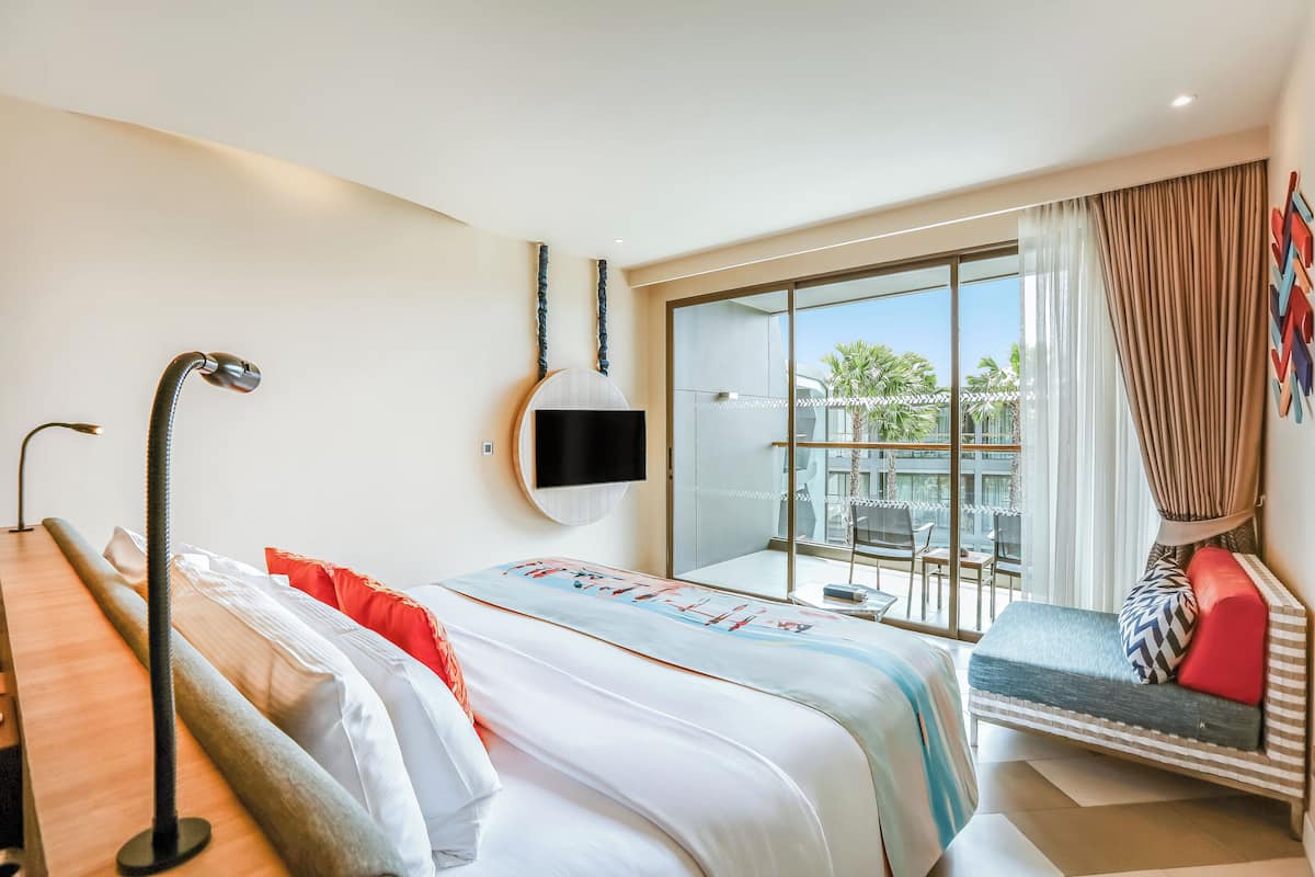 Habitación de hotel con cama, escritorio, televisor y balcón con vista a palmeras.