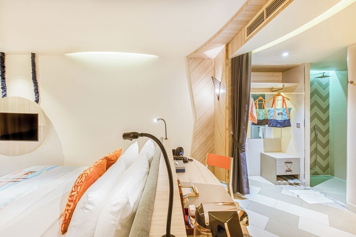 A modern hotel room with a bed, a desk, a chair, a desk lamp, a mirror, a TV, a shower area, and a bathroom with a towel rack.