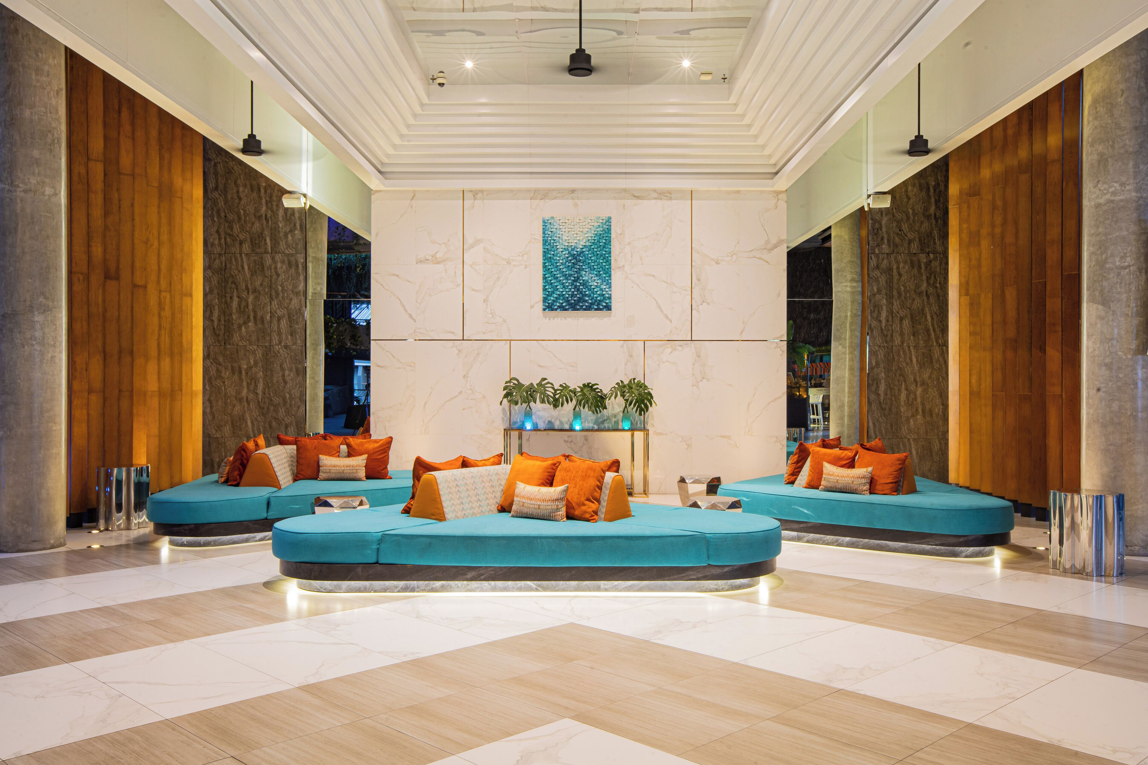 Eine großzügige Lobby mit einer zentralen Sitzecke, ausgestattet mit türkisfarbenen Sofas und orangefarbenen Kissen, umgeben von Marmorwänden und Holzelementen.
