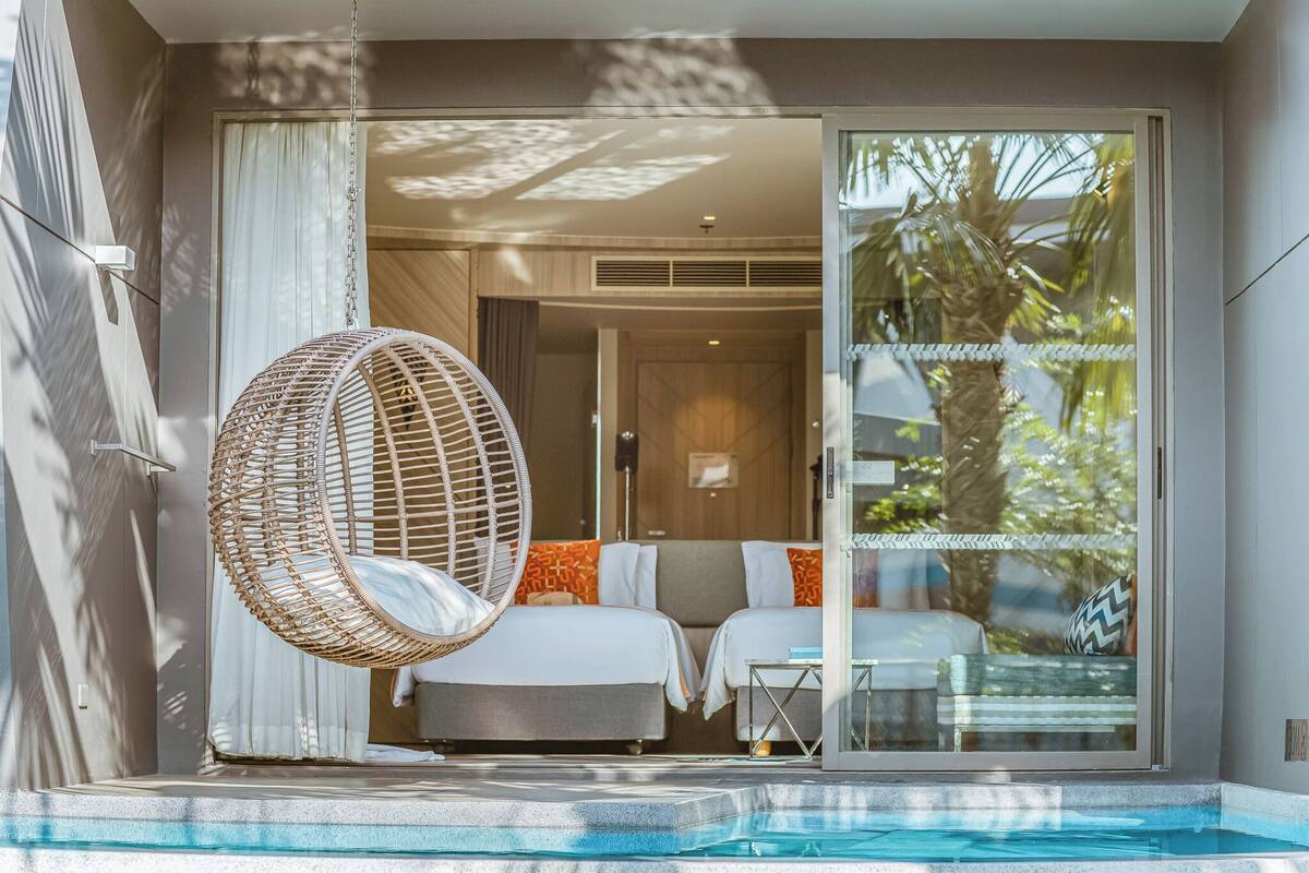Una habitación moderna con puerta de vidrio que da a una piscina, un sillón colgante y dos camas con almohadas.