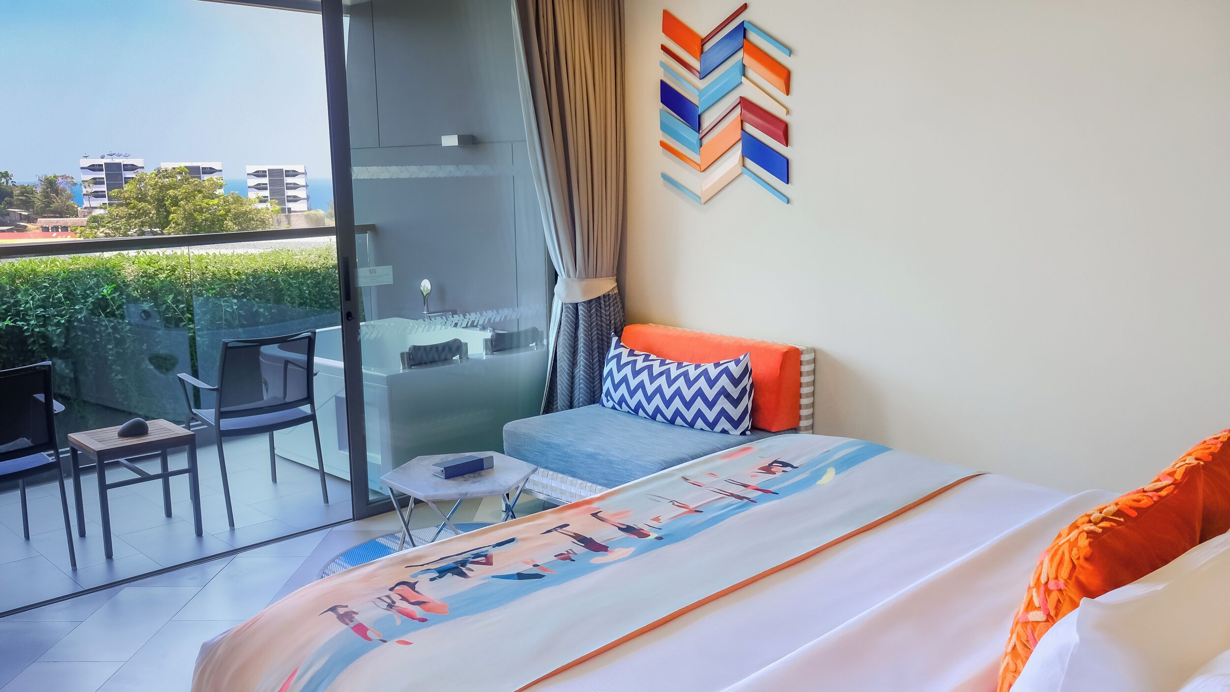 Una habitación de hotel moderna con balcón, cama con colcha colorida, una mesita y una silla.