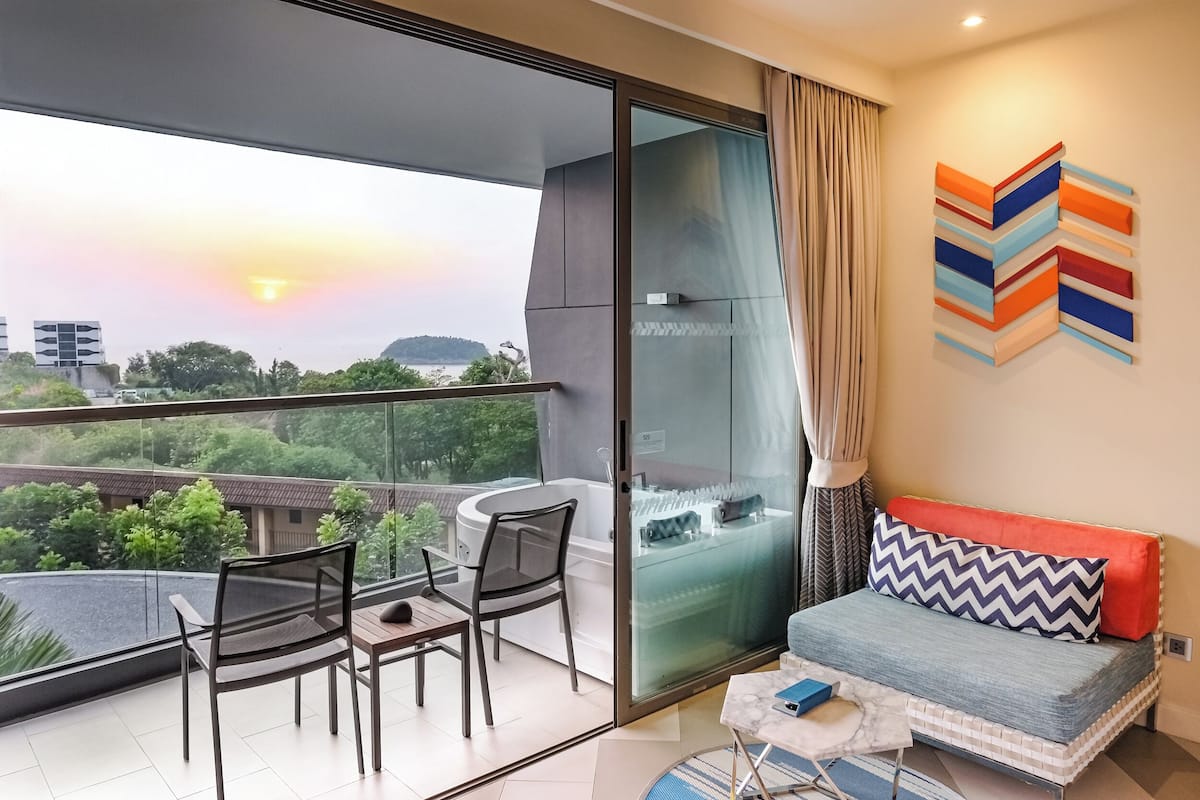 Un balcón con vistas a un atardecer, un sofá con cojines estampados y una puerta de vidrio que da a un baño.