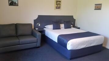 Quarto de hotel com uma cama, um sofá cinzento, uma mesinha de cabeceira e uma luminária fixada na parede.
