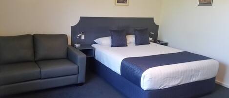 A hotel room with a bed, a grey sofa, a nightstand, and a wall-mounted lamp.
