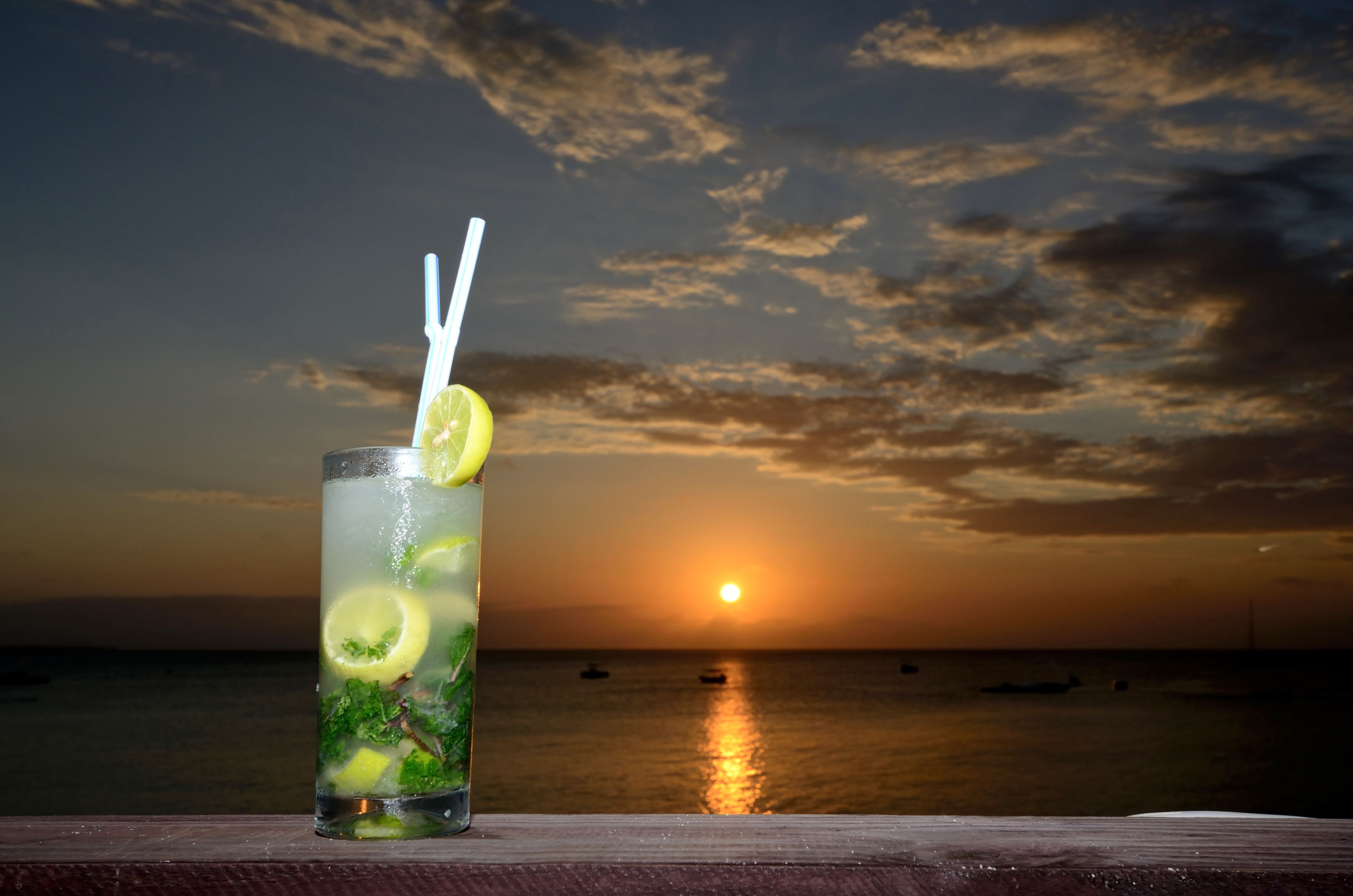 Un verre de boisson glacée, avec une paille et une tranche de citron, posé sur une surface en bois, avec en arrière-plan un coucher de soleil sur l’océan.