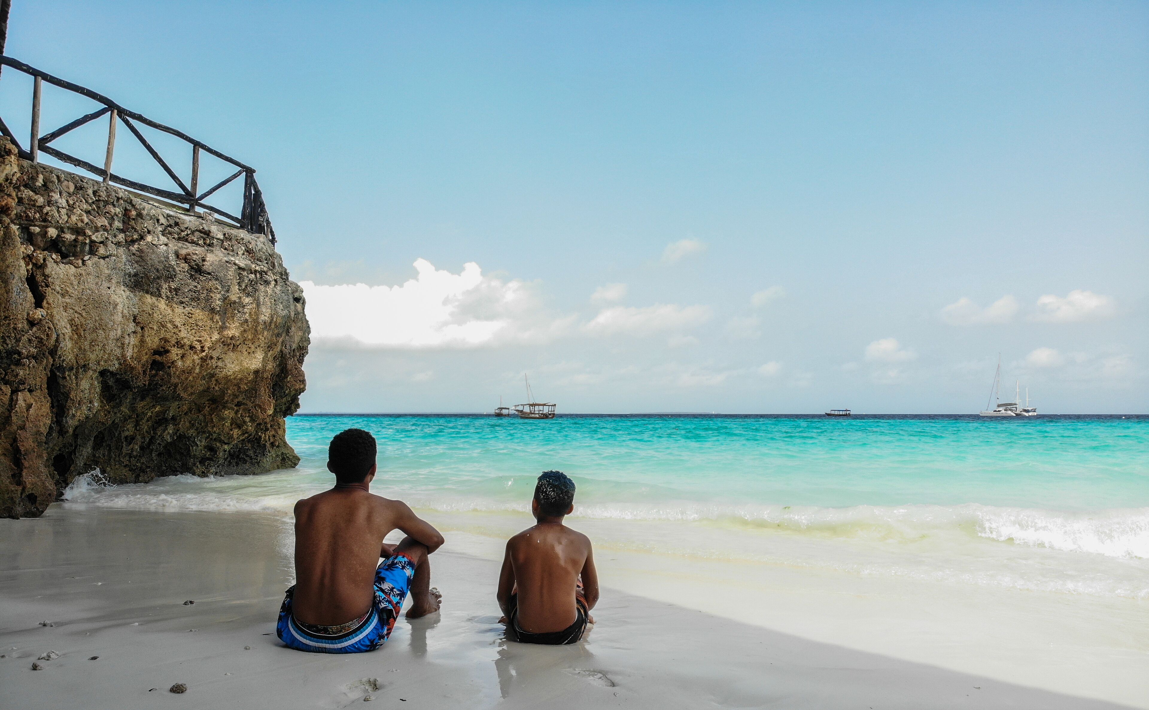 Dos personas sentadas en una playa arenosa con un acantilado rocoso y el océano al fondo.