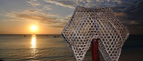 A beach scene with a netted structure, a wooden post, and the ocean with boats in the distance during sunset.