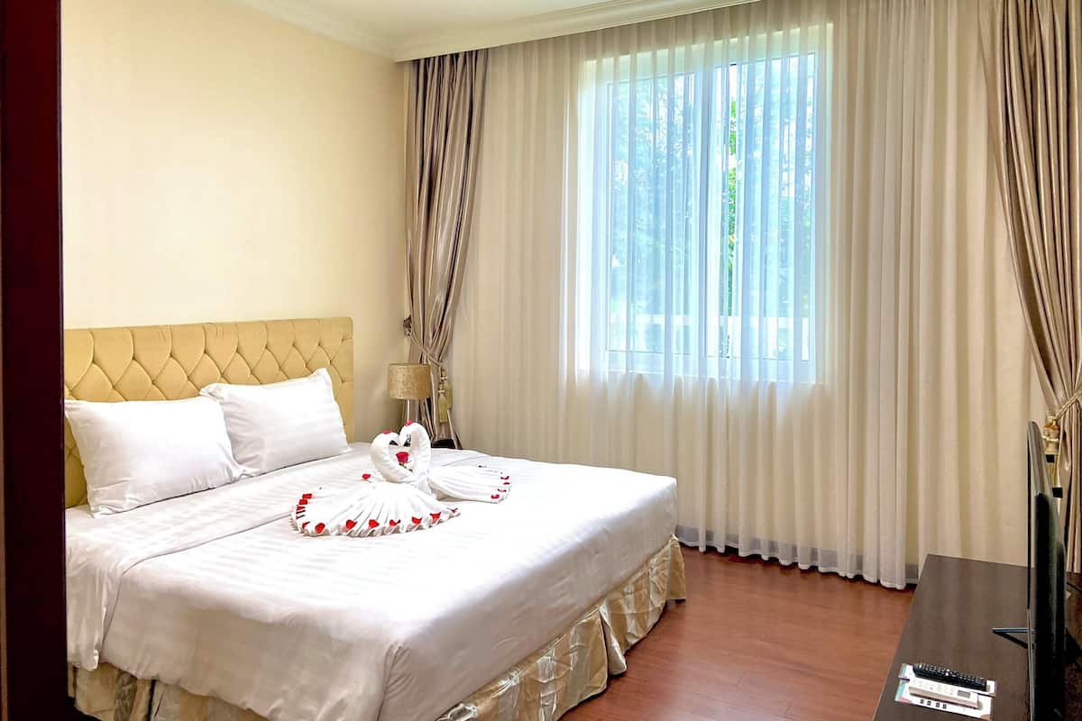 A neatly made bed with white linens and decorative pillows, a wooden headboard, and a large window with sheer curtains.
