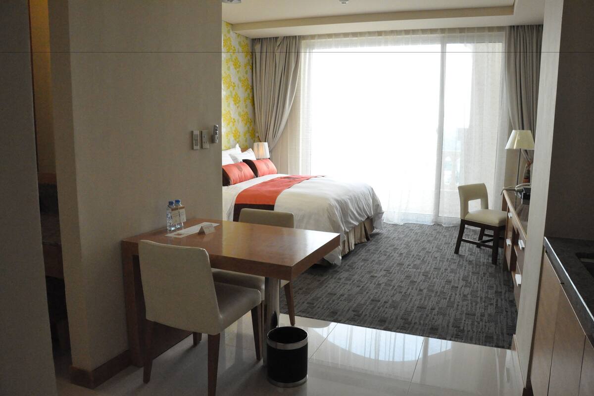 A hotel room with a bed, a dining table, chairs, a small stool, a trash can, and a window with curtains.