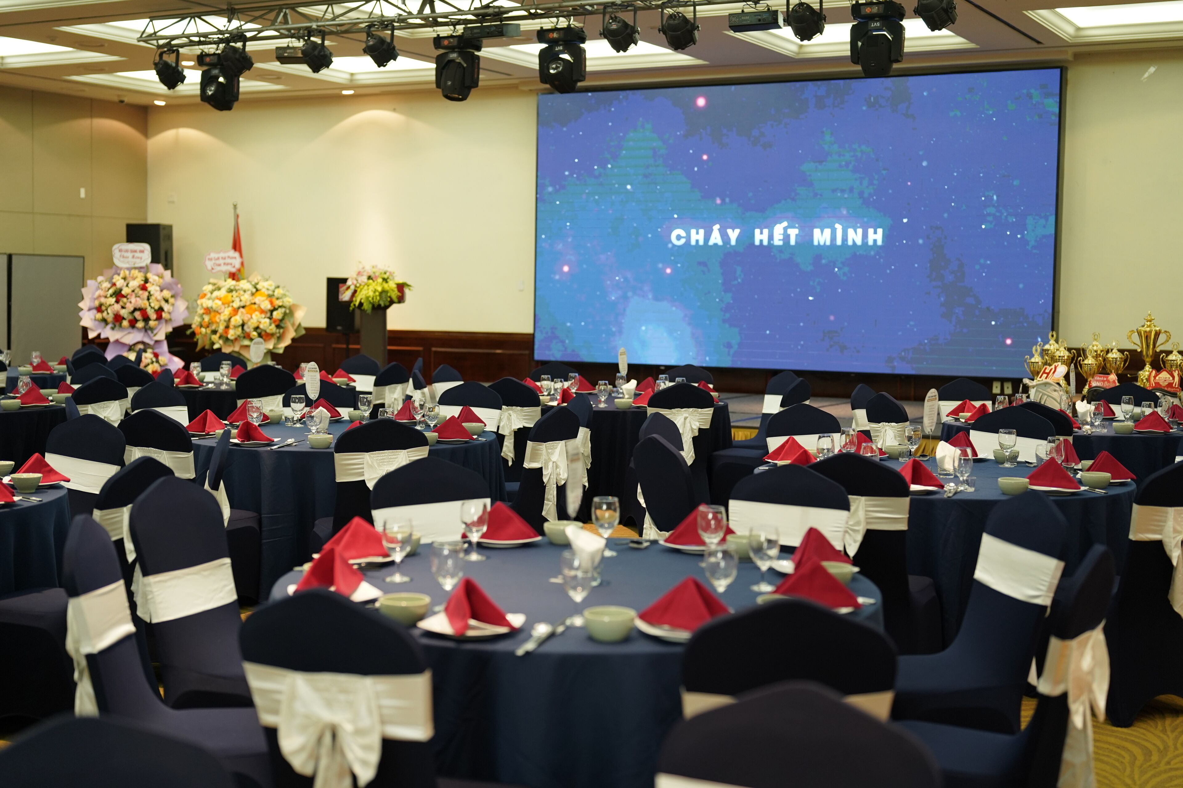 A banquet hall with round tables set for a formal event, featuring navy chairs with white sashes, red napkins, and a large screen displaying text.