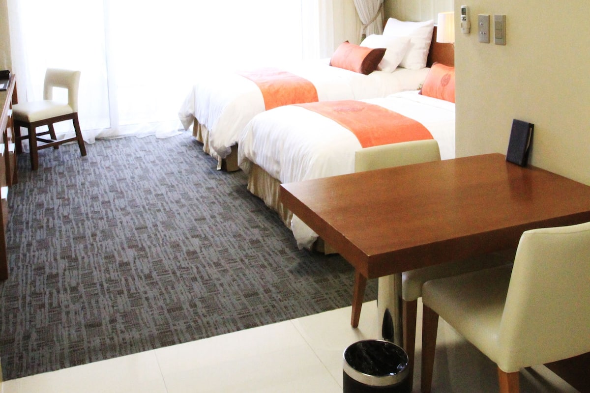 A hotel room with a bed, a desk, a chair, a table, a trash can, and a picture on the wall.