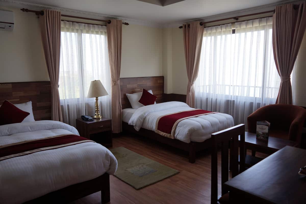 Quarto de hotel com duas camas, cabeceira de madeira, mesa de cabeceira com abajur, uma cadeira e uma pequena mesa com um livro.