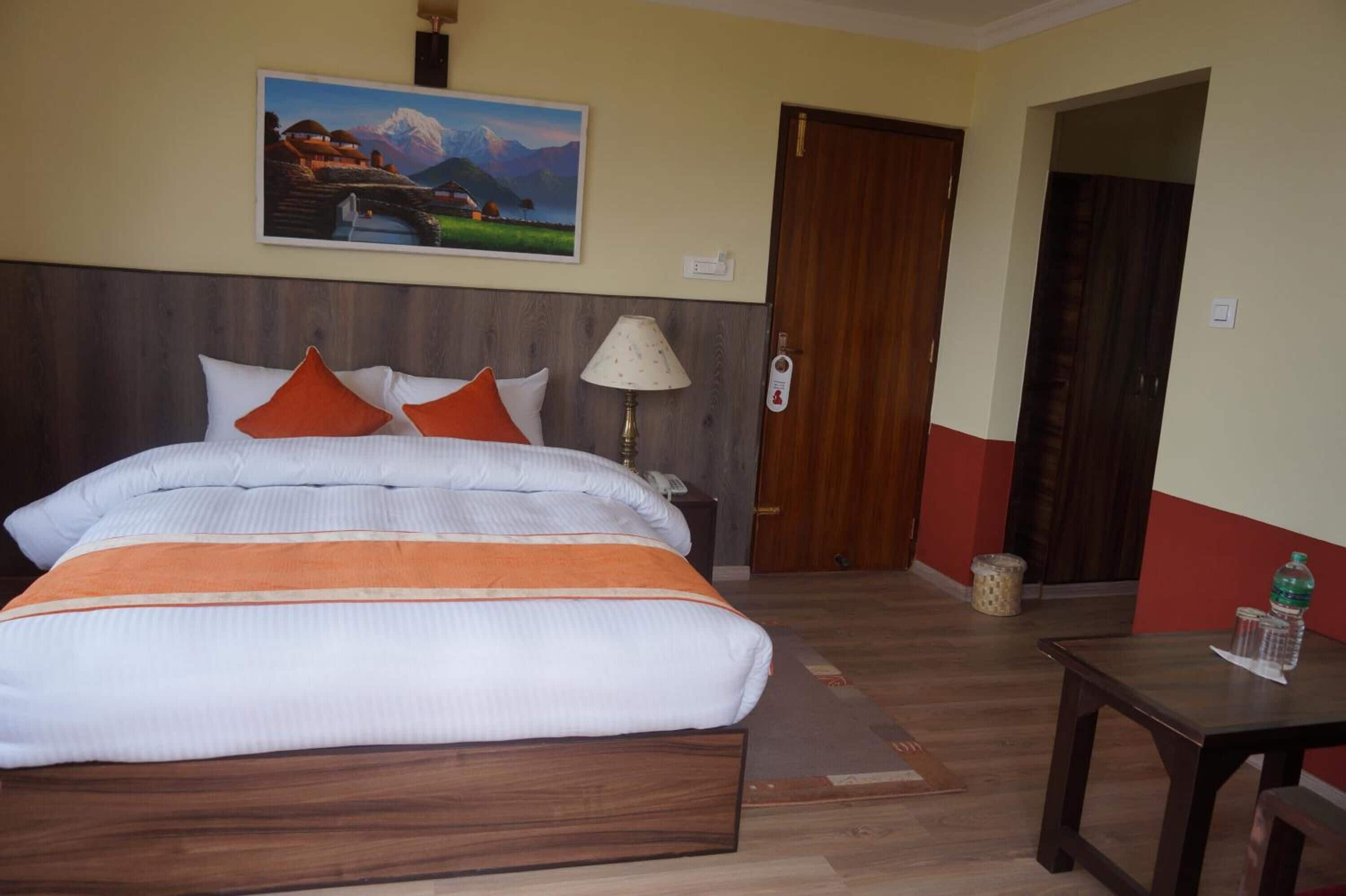 A hotel room with a large bed, a painting on the wall, a bedside lamp, a wooden nightstand, and a small table with a bottle and glass.