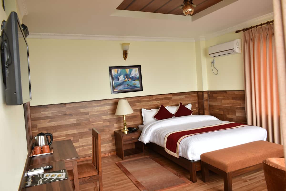 Quarto de hotel com cabeceira de madeira, cama com roupa de cama branca e almofadas vermelhas, mesa de cabeceira com luminária, pequena escrivaninha com cafeteira e um quadro na parede.