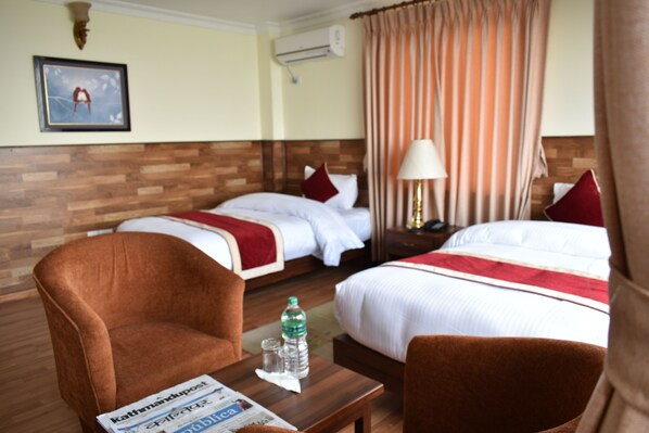 Habitación de hotel con dos camas, cabecero de madera, sillón marrón, una mesa con un periódico, una botella de agua y una lámpara.