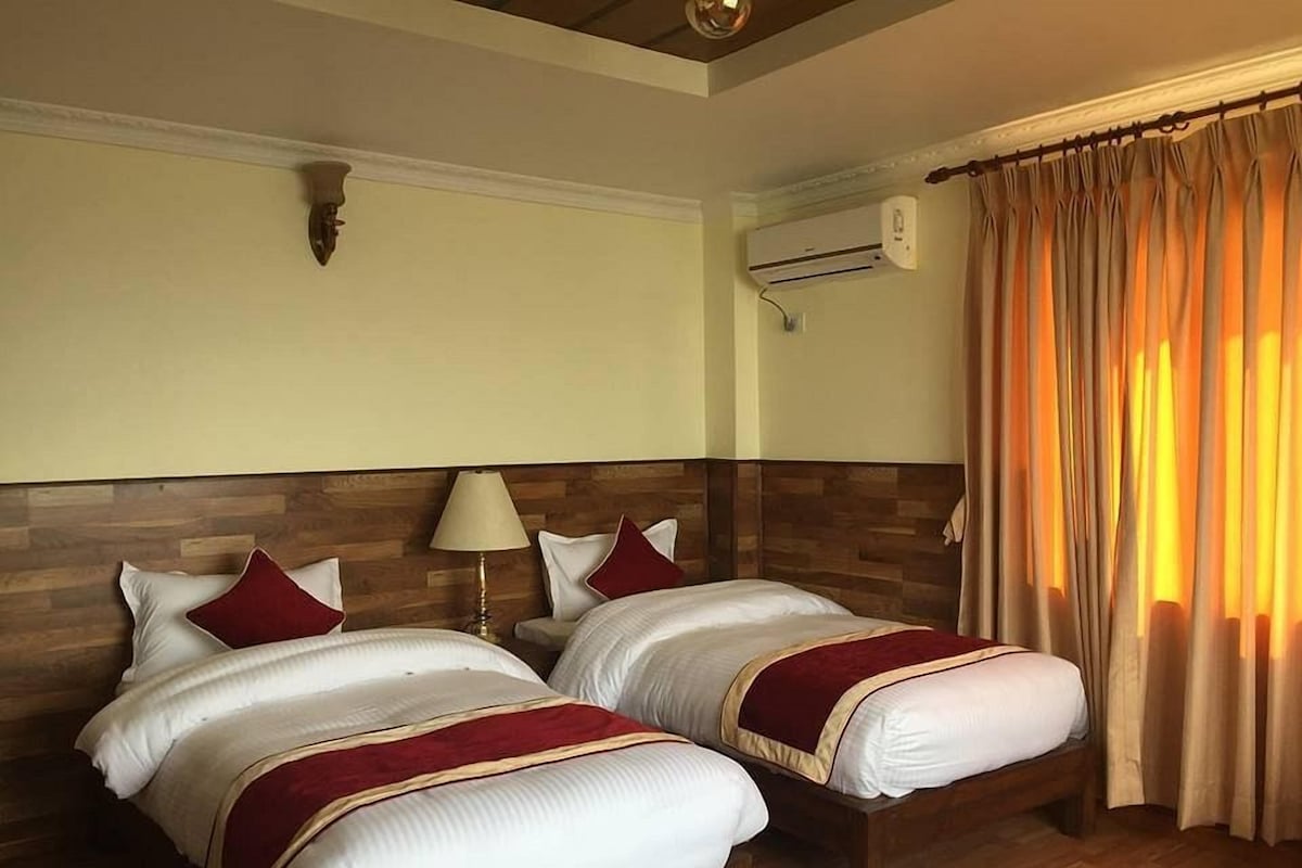 A hotel room with two beds, a wooden headboard, a nightstand with a lamp, an air conditioning unit, and a window with curtains.