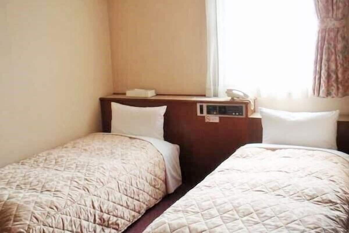 Une chambre d’hôtel avec deux lits simples, une table de chevet avec un téléphone et une lampe, et une fenêtre avec des rideaux.