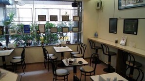 A restaurant with multiple tables and chairs, a window with framed pictures, and a view of greenery outside.