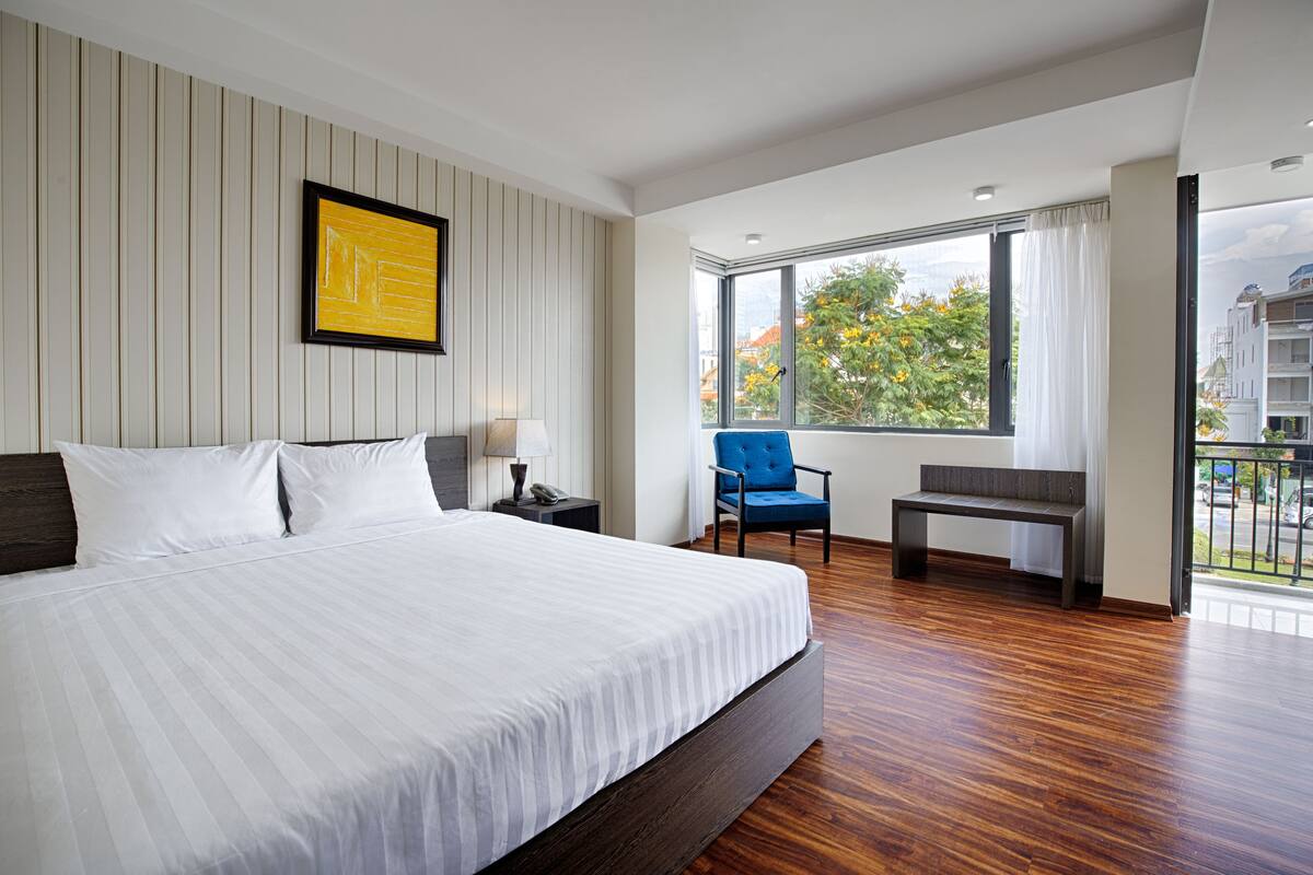 Une chambre d’hôtel moderne avec un grand lit, une chaise bleue, une petite table et une vue sur la ville grâce à de grandes fenêtres.