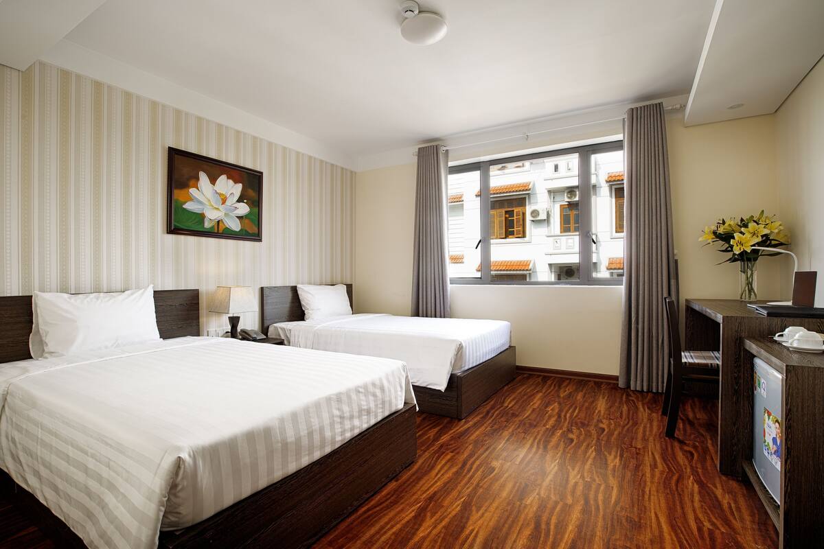 Une chambre d’hôtel avec deux lits, un bureau, une chaise, une fenêtre avec des rideaux et un tableau au mur.
