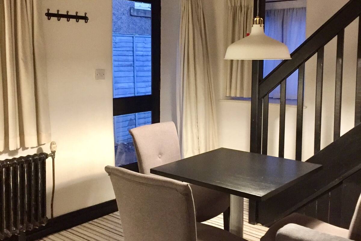 Un coin repas avec une table et des chaises, des poutres en bois au plafond, une fenêtre avec des rideaux et un escalier.