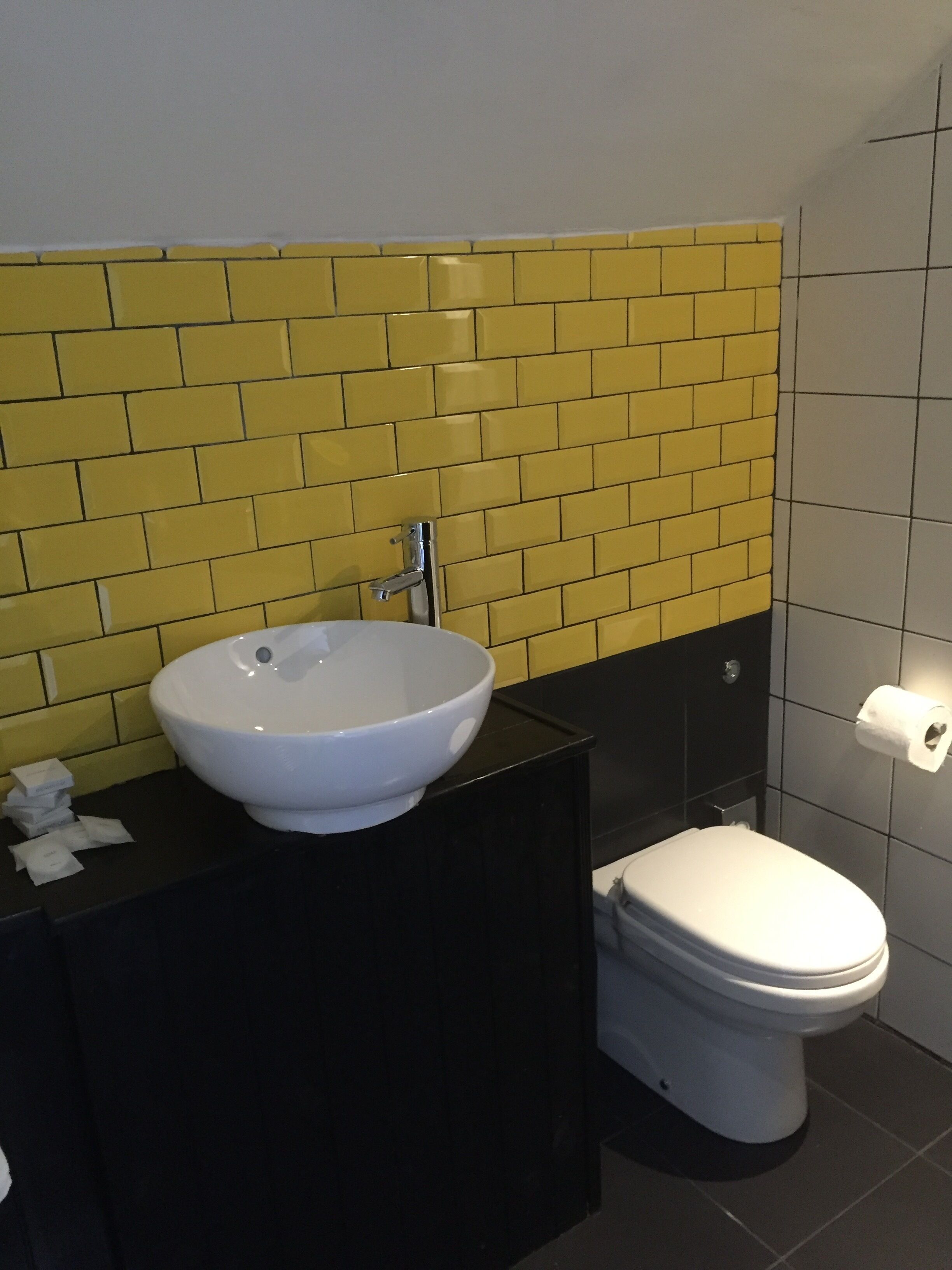 A bathroom with a white sink, black countertop, and a toilet. The walls are tiled with yellow and white tiles.