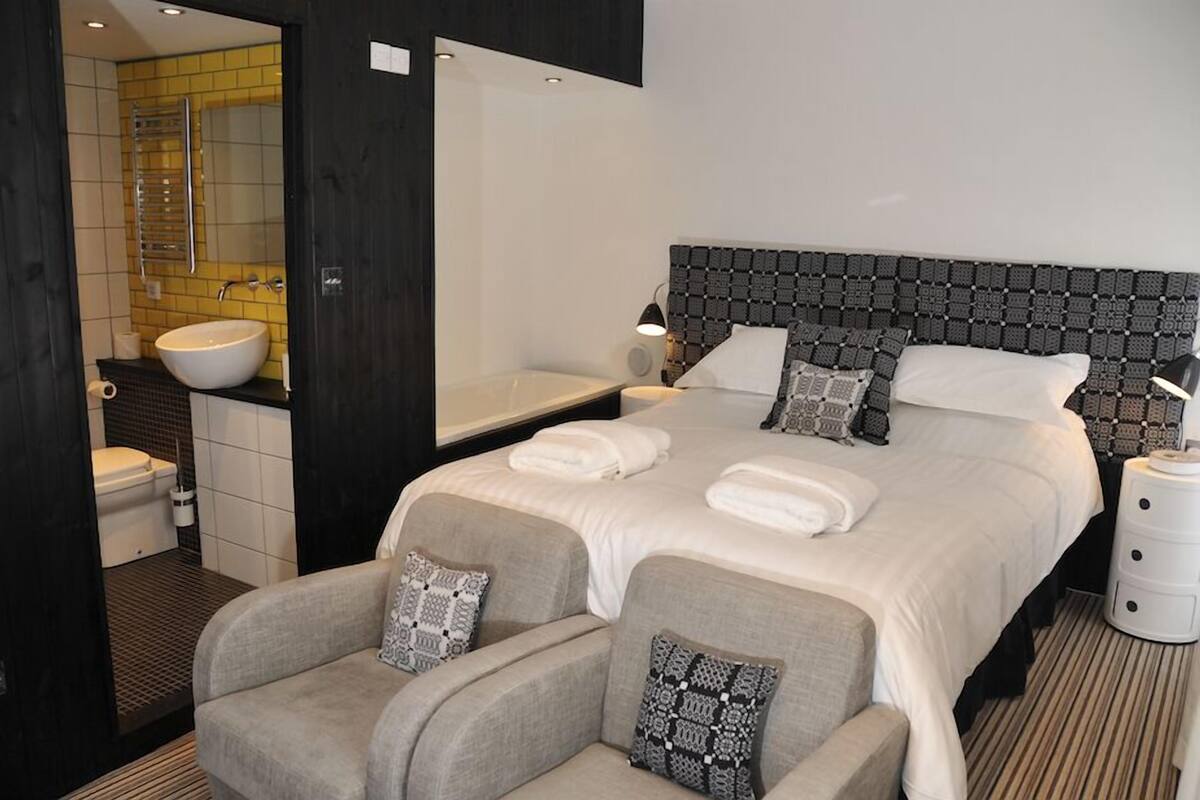 Une chambre d’hôtel moderne, dotée d’un grand lit, de deux fauteuils, d’une table de chevet, d’une lampe et d’une salle de bain avec un lavabo et des toilettes.