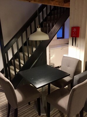 Un escalier avec une rampe noire, un luminaire suspendu blanc et un petit coin cuisine avec un meuble blanc et une couverture anti-feu rouge.