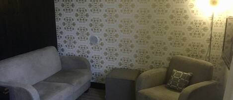Ein Zimmer mit geprägtem Wandpapier, einem grauen Sofa, einem kleineren Sessel und einer Stehlampe.