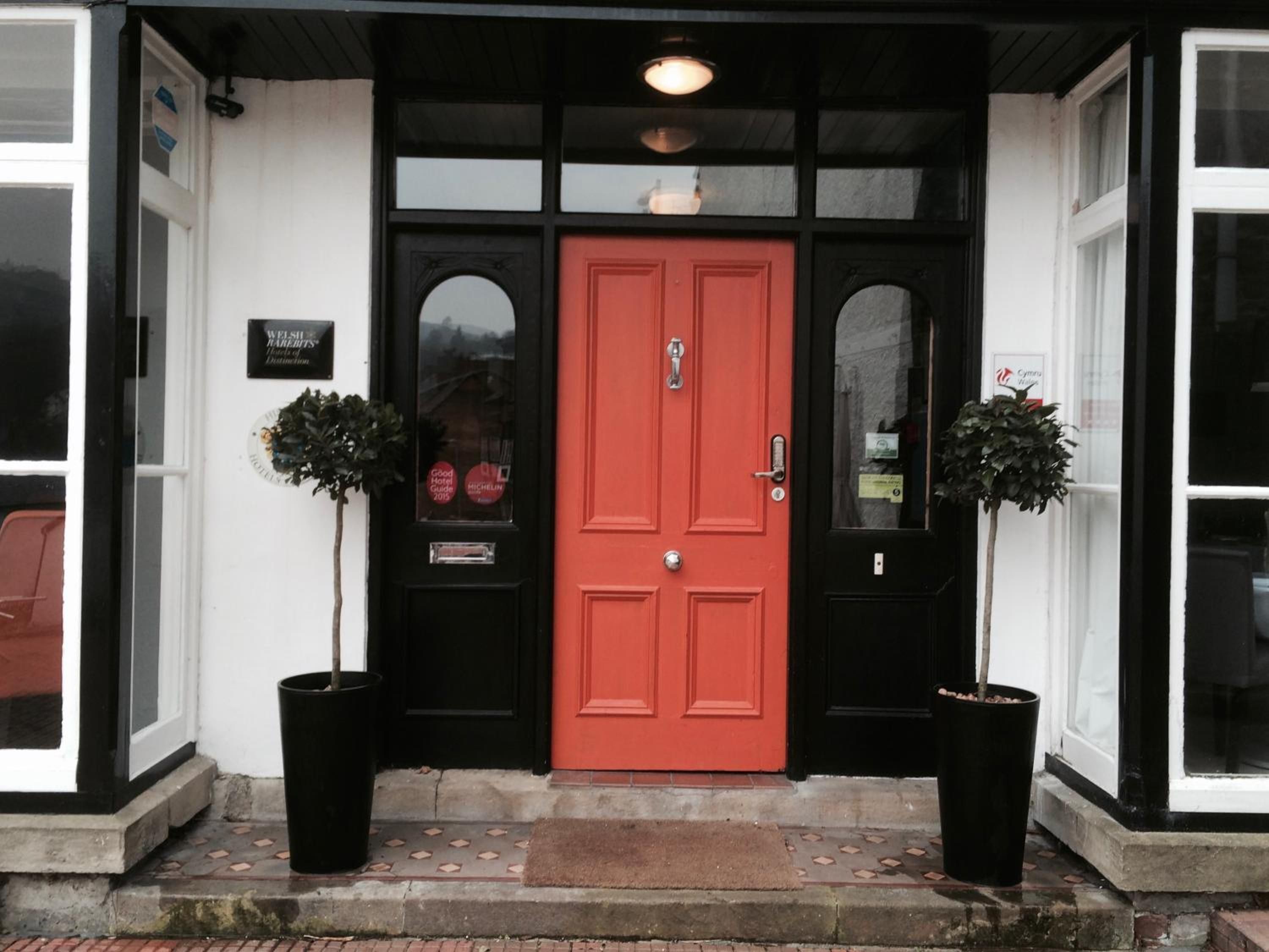 L’entrée d’un hôtel, avec une porte rouge, des fenêtres à cadre noir et deux plantes en pot de chaque côté.