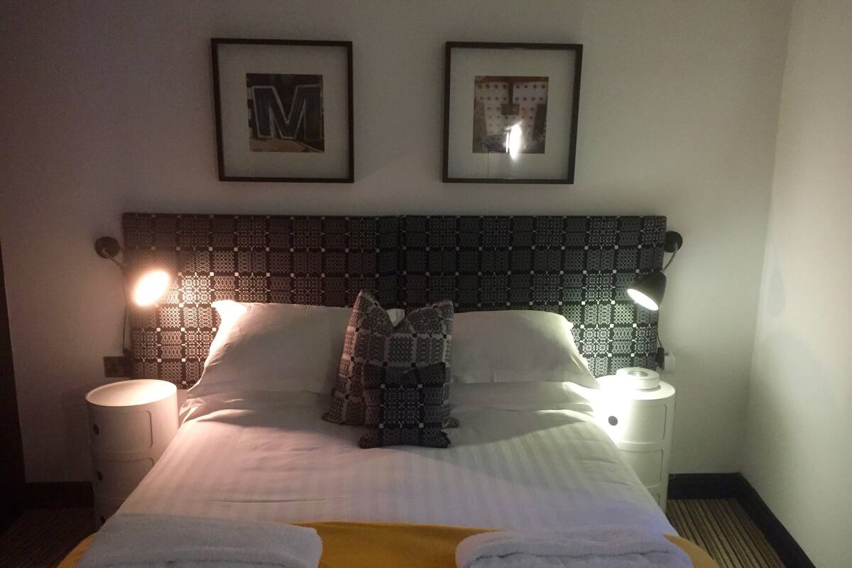 Un lit bien fait, avec du linge de lit blanc, deux oreillers et une couverture jaune. On y trouve deux lampes de chevet, une lettre « M » encadrée accrochée au mur et un tableau encadré au-dessus du lit.