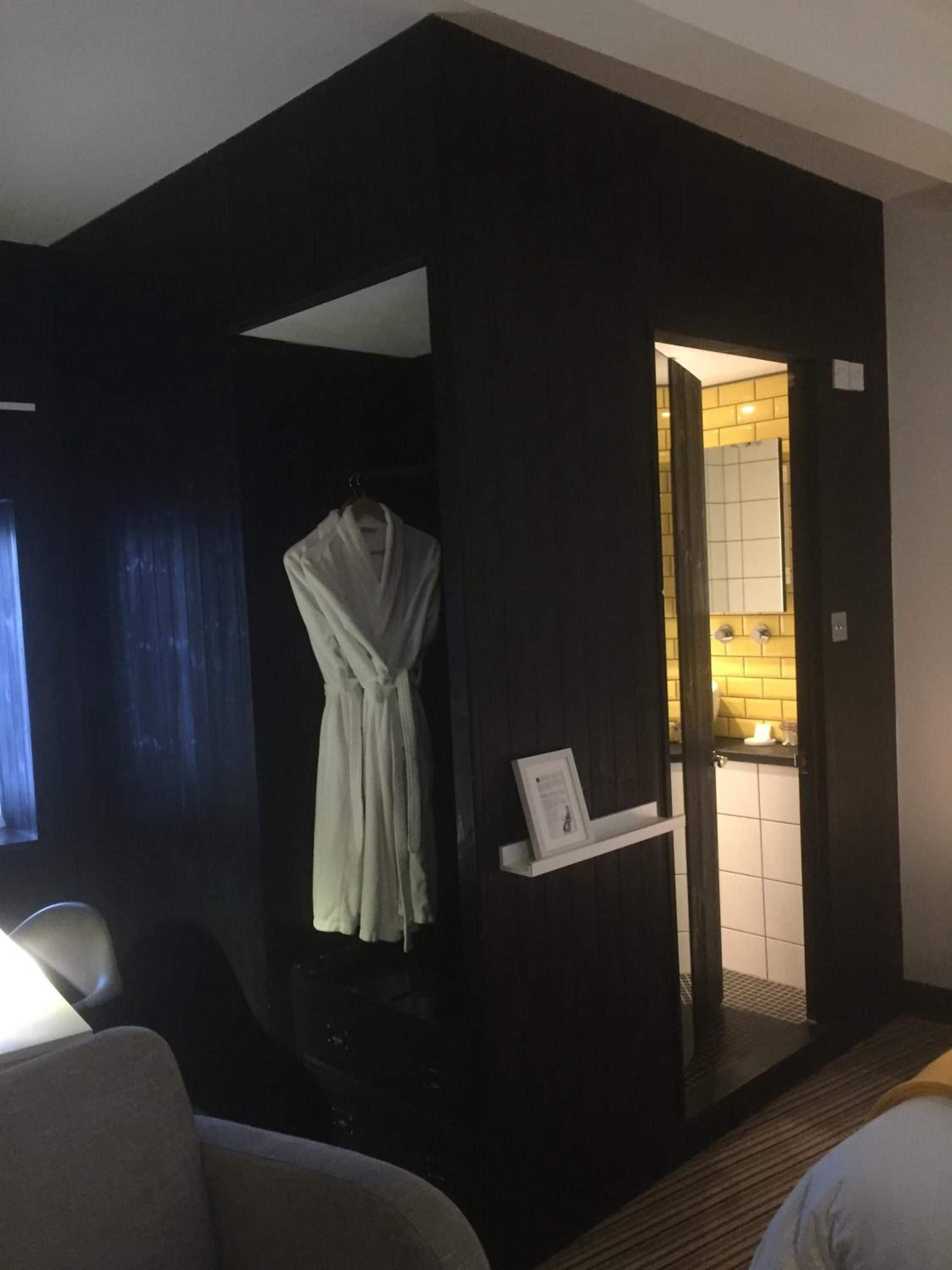 A hotel room with a dark-colored wall, a white robe hanging on a hook, a small white shelf with a framed picture, and a glimpse of a bathroom with a yellow-tiled wall.