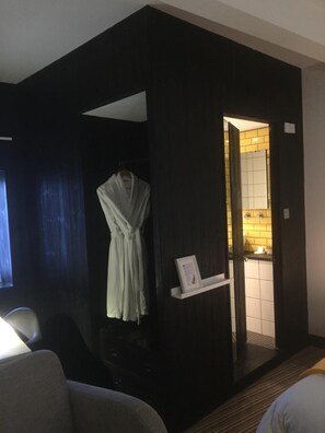 A hotel room with a dark-colored wall, a white robe hanging on a hook, a small white shelf with a framed picture, and a glimpse of a bathroom with a yellow-tiled wall. - Greenbank Lodge Llangollen (Llangollen)