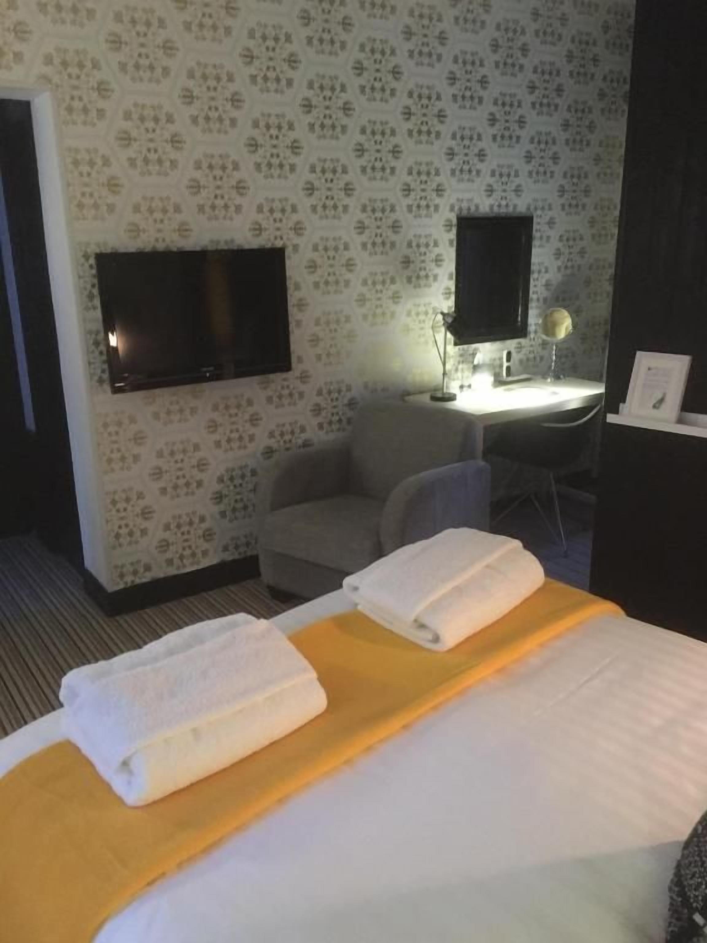Een hotelkamer met een bed, een bureau met een lamp, een aan de muur bevestigde televisie en behang met een patroon.