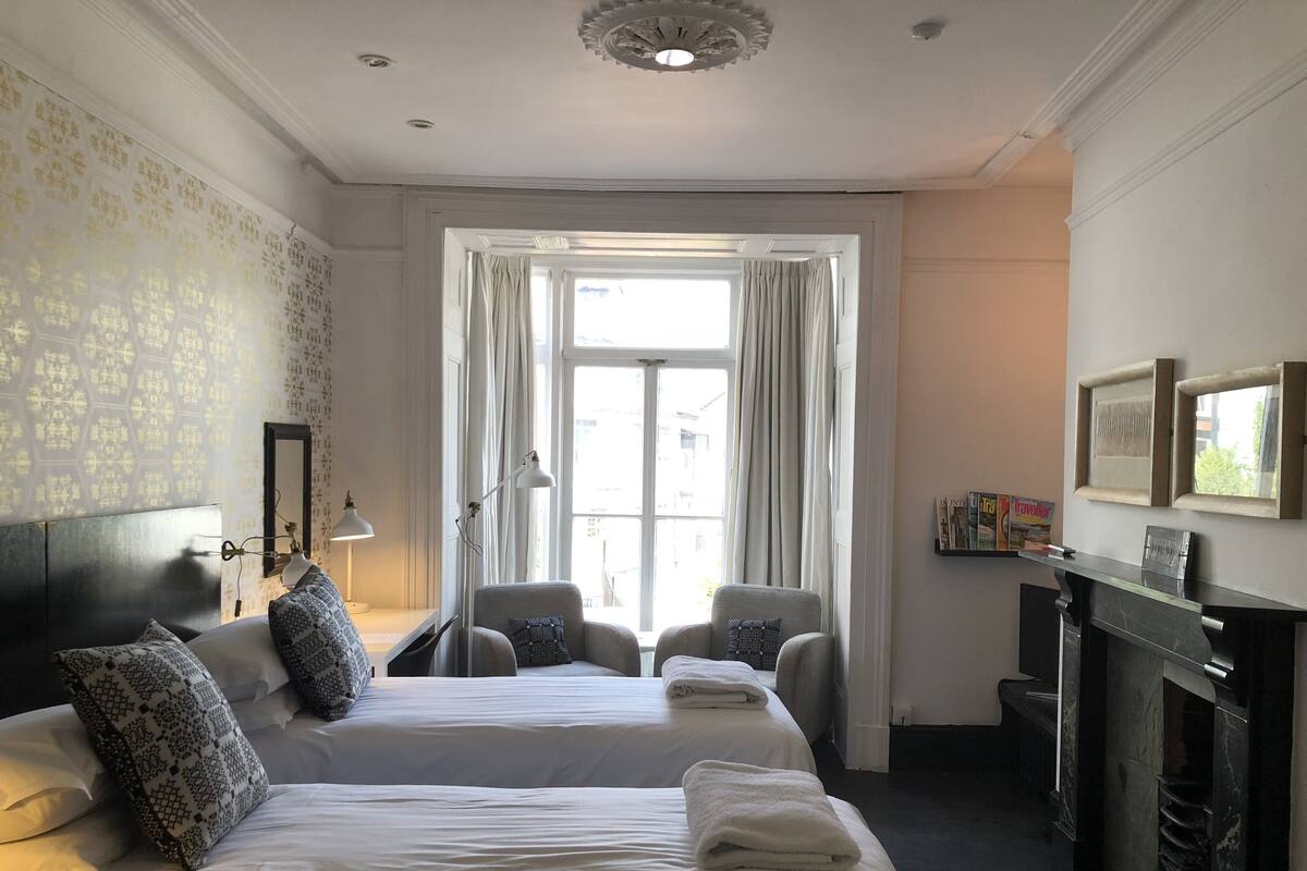 Une chambre d’hôtel avec deux lits, un coin salon avec des fauteuils, une cheminée et une grande fenêtre avec des rideaux.