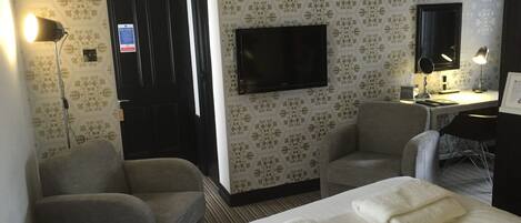 A hotel room with two chairs, a bed with towels, a desk with a computer, a TV, and a patterned wallpaper.